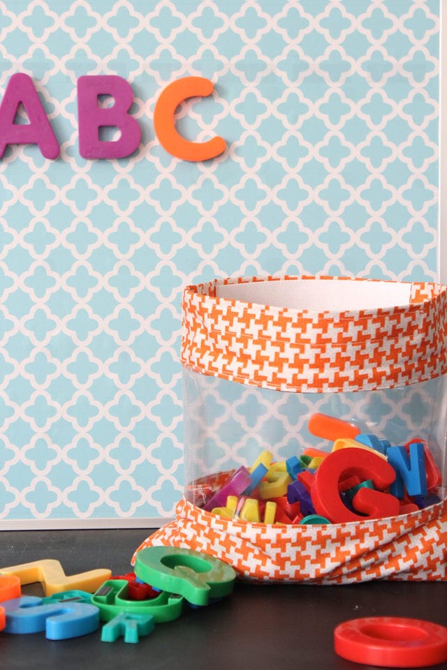 An orange houndstooth fabric toy bag with a clear vinyl window full of colorful magnetic letters against a blue wall
