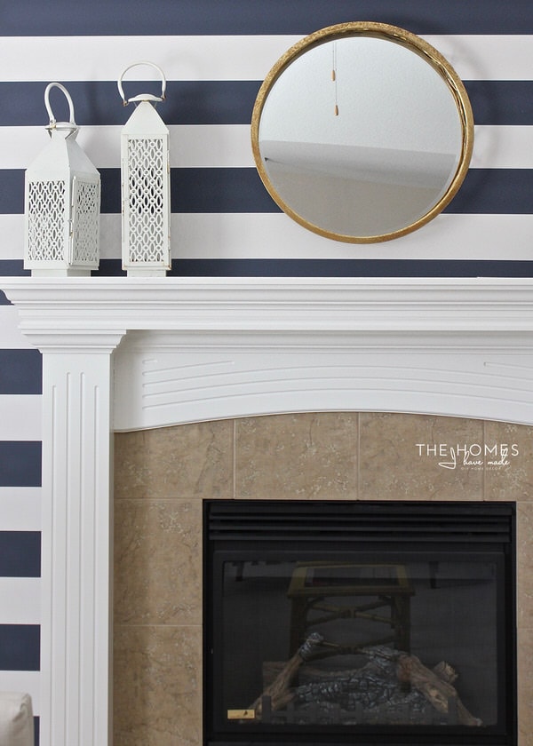 a photo of a fireplace surround with navy and white wall papered walls, a round, gold mirror and white lanterns on the mantel.