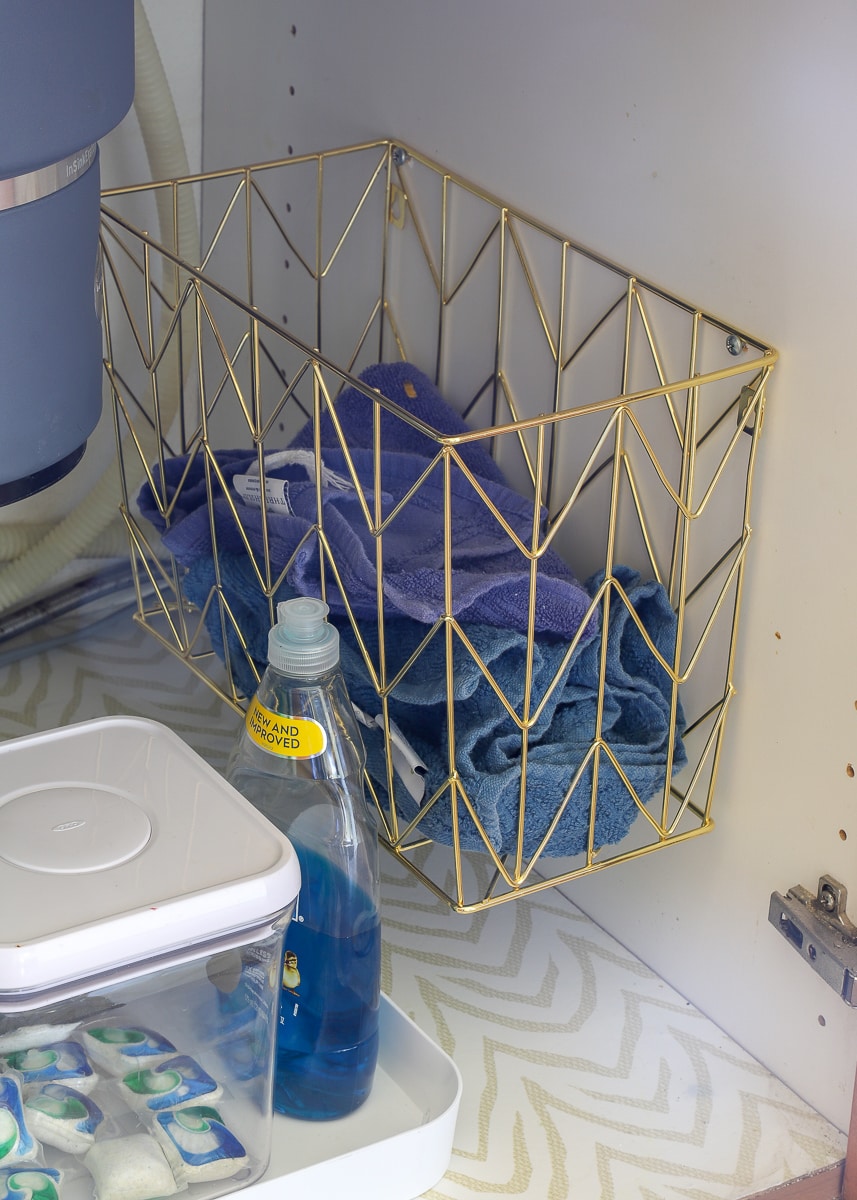 A gold wired hanging file basket under the sink containing dirty dish towels