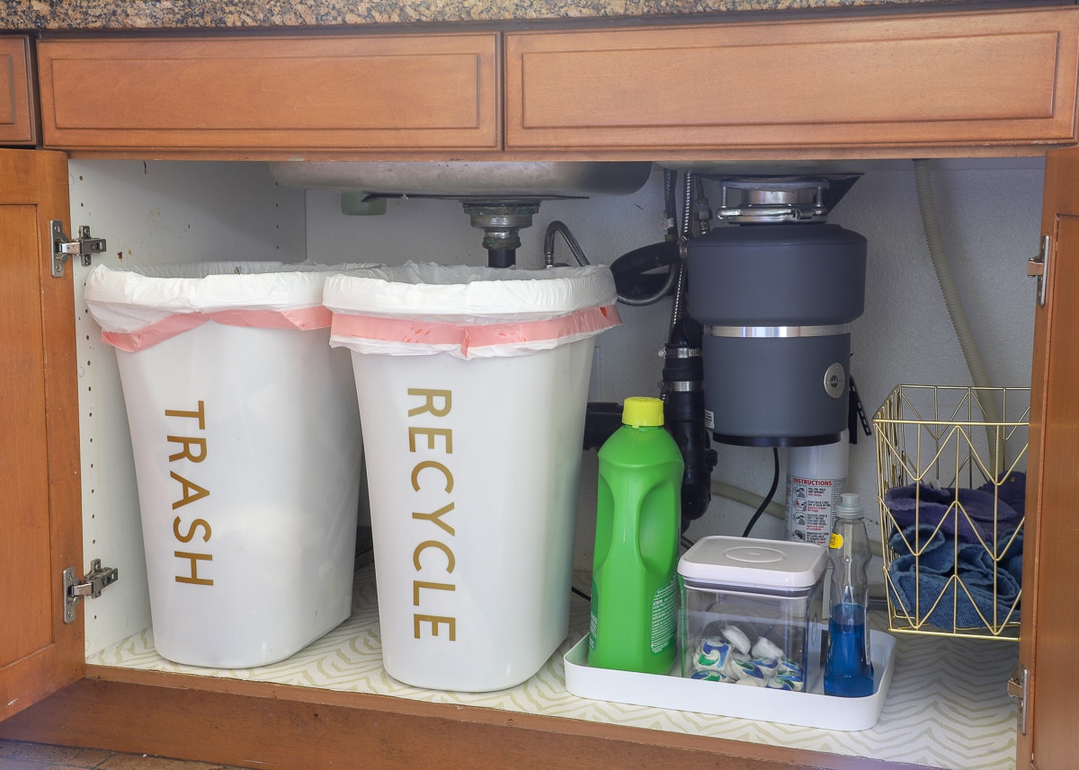 Dirty dish towels, dish washing supplies, and trash cans organized in a cabinet under the sink