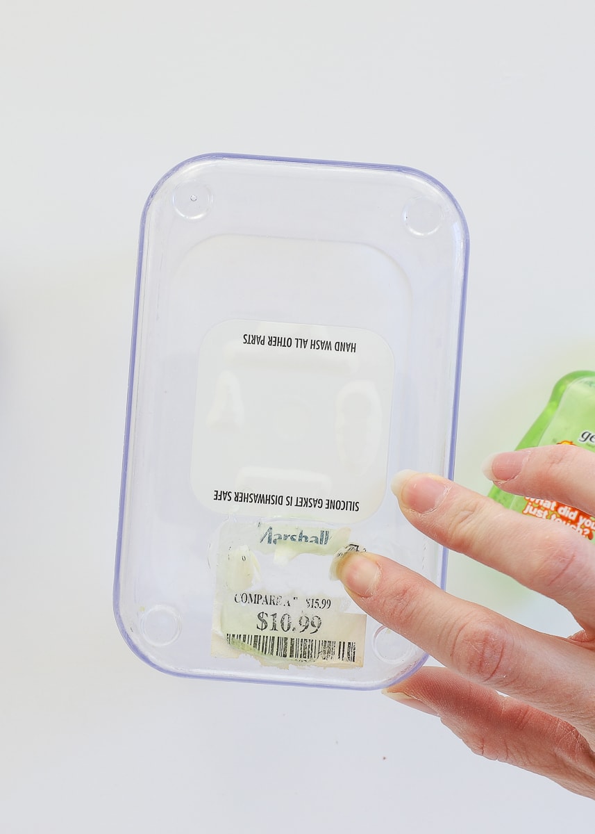 Finger nail scraping up a white paper label off a food canister after covering it in hand sanitizer to remove glue
