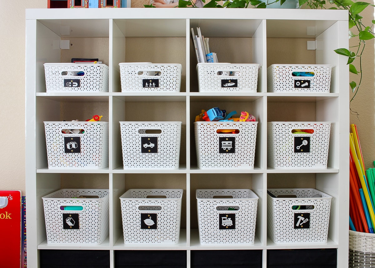 White baskets with black labels filled with toys on an IKEA Kallax shelf