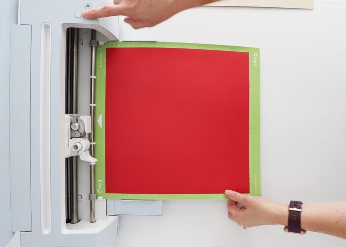 Hand loading a green Cricut mat with red paper into a Cricut Maker 3 machine.