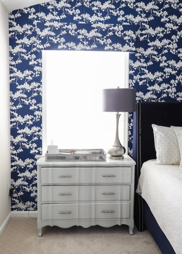 Rental bedroom with blue and white flowered wallpaper with a silver nightstand and a grey lamp.