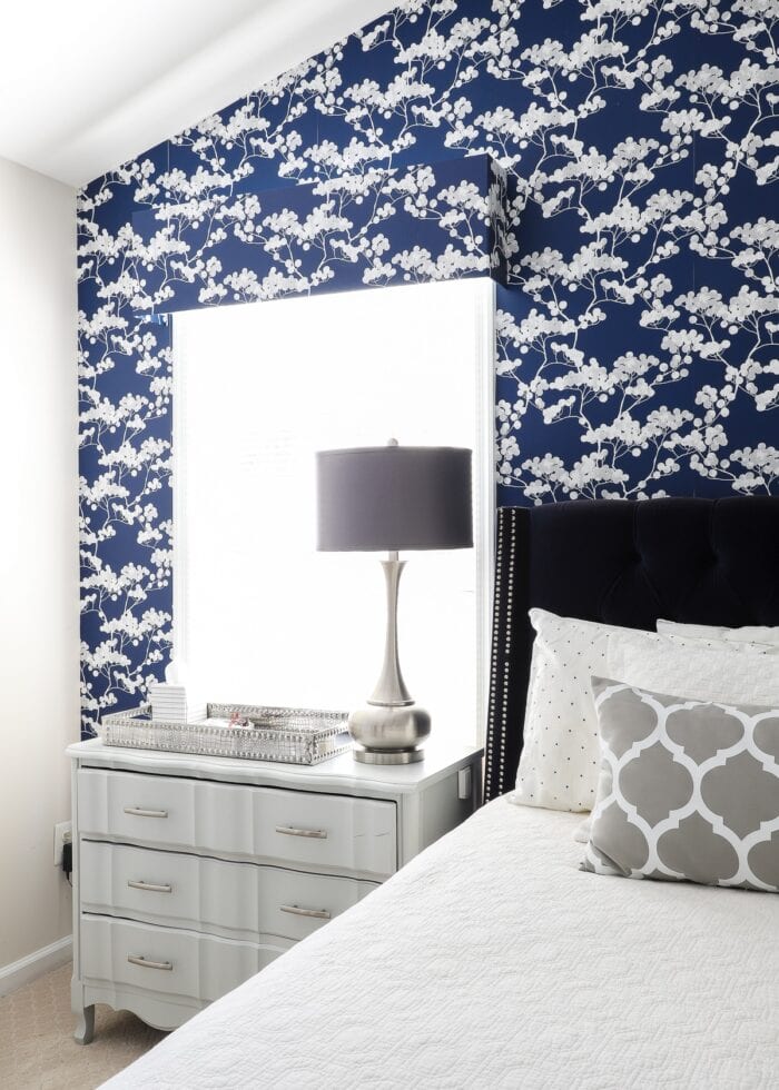 Rental bedroom with blue and white flowered wallpaper with a silver nightstand and a grey lamp.