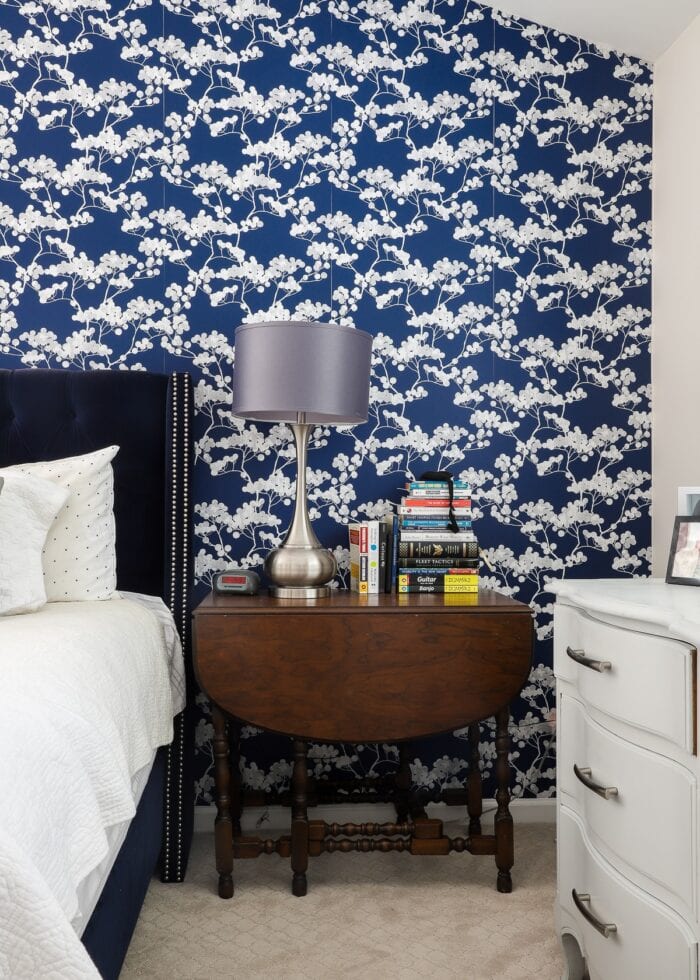 Rental bedroom with blue and white flowered wallpaper a vintage wood side table, and a grey lamp.