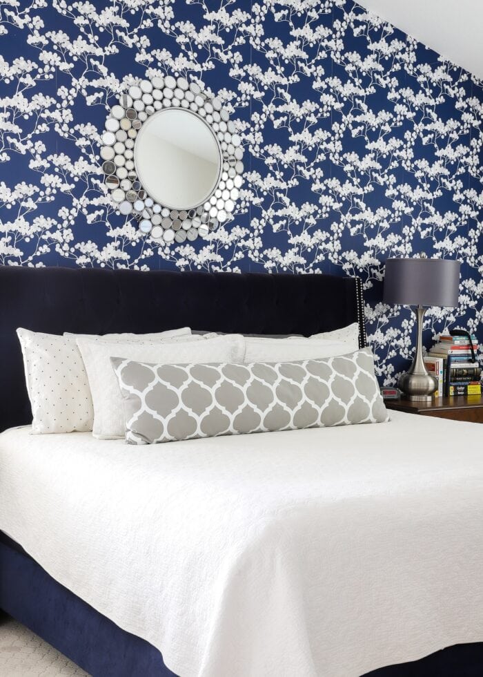 Rental bedroom with blue and white flowered wallpaper, a navy bed with a white bedspread and grey lamps.