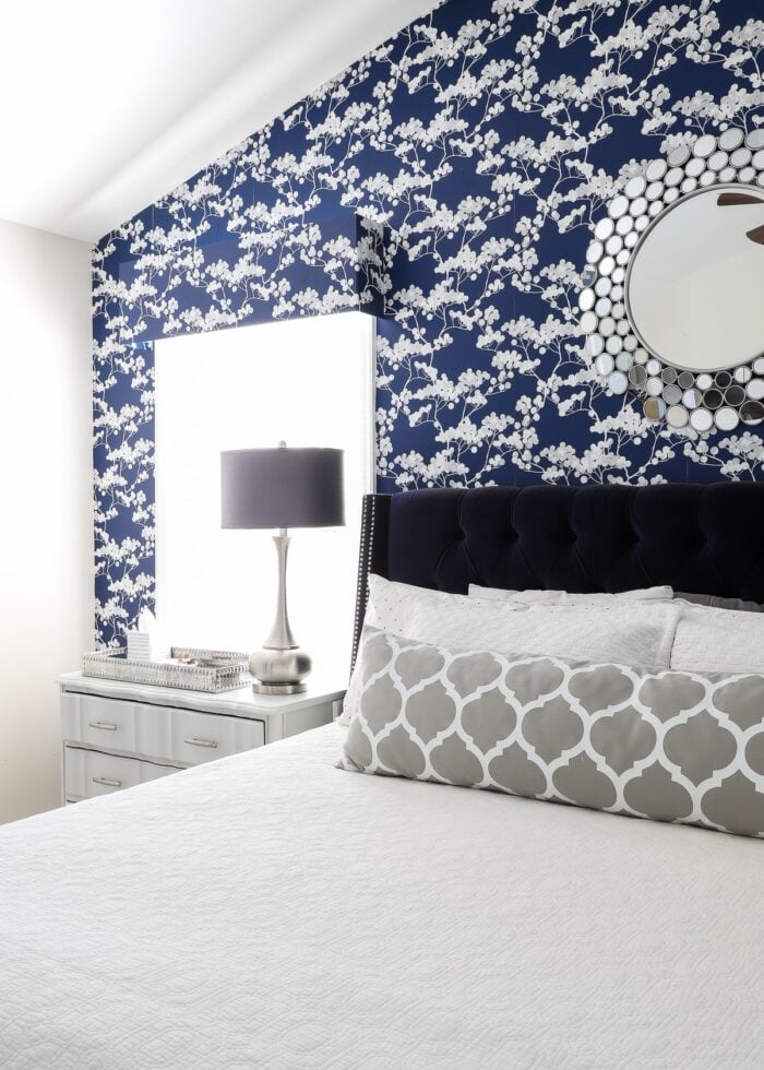 Rental bedroom with blue and white flowered wallpaper, a navy bed with a white bedspread and grey lamps.