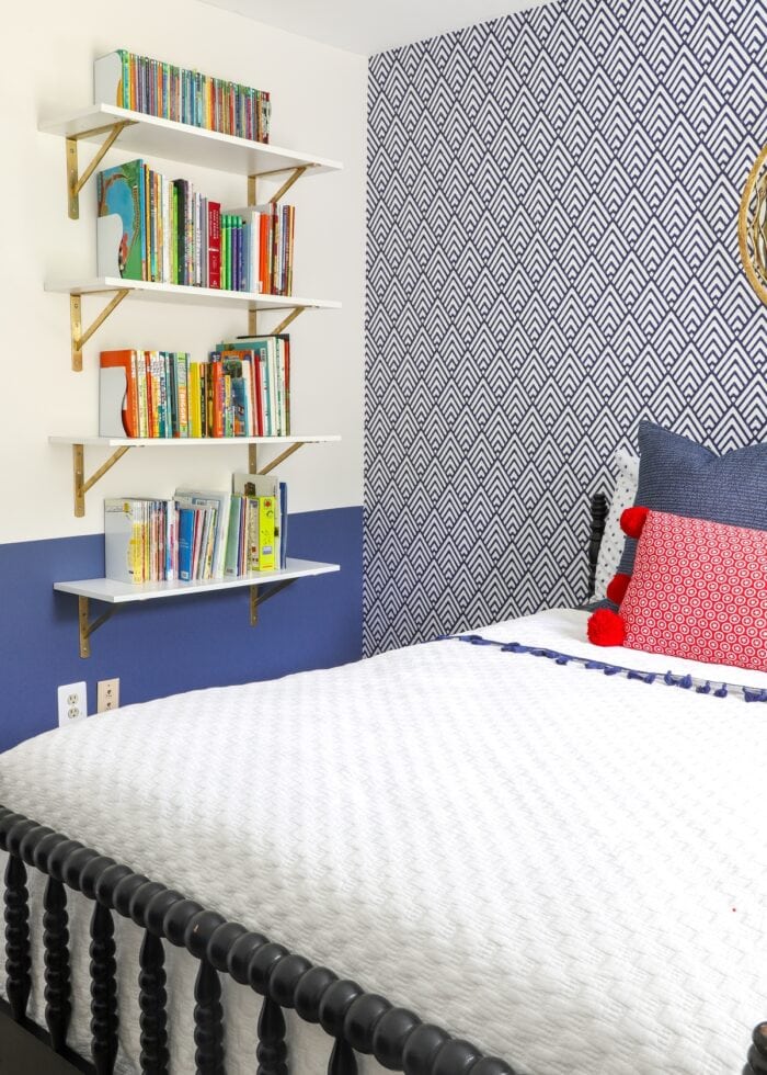 Blue and white wallpaper behind a black bed with red, white, and blue linens and a bookshelf on the wall