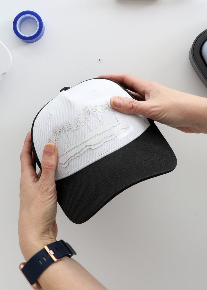 Hands placing an Infusible Ink design onto the front of a black trucker hat.