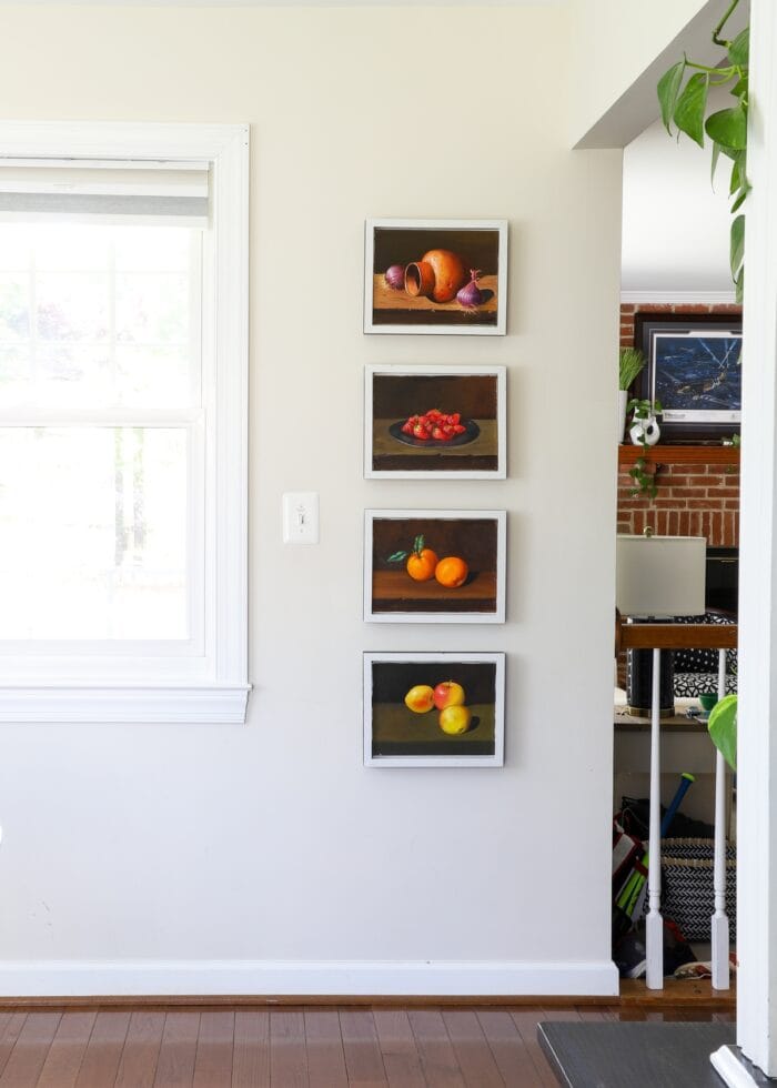 A neutral wall with four framed oil paintings of fruits and vegetables hung in a vertical arrangement