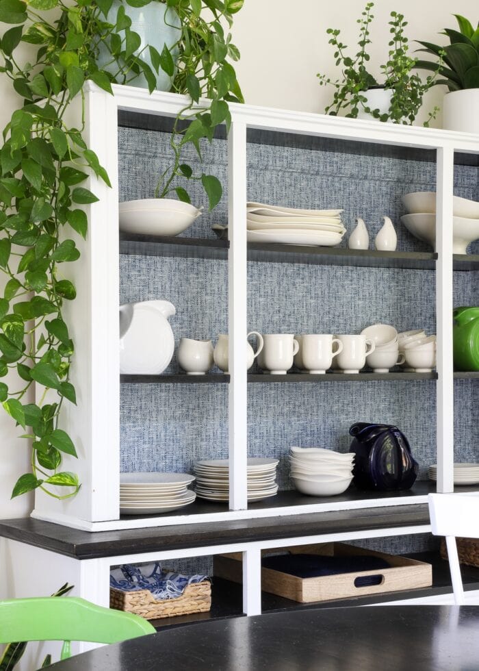 A white China Hutch with blue wallpaper along the back, white dishes, and green plants on top