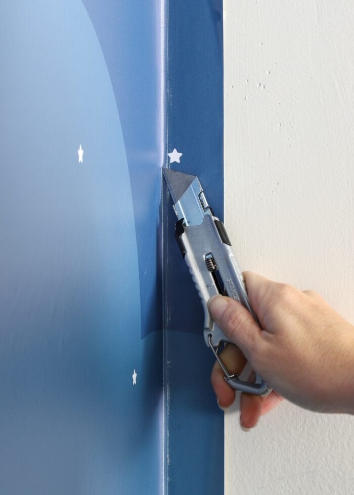 Hand using sharp blade to cut away excess wallpaper mural