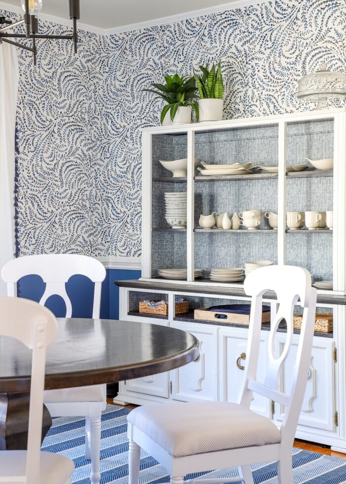 White china hutch in a formal dining room with blue and white wallpaper on the walls above chair rail