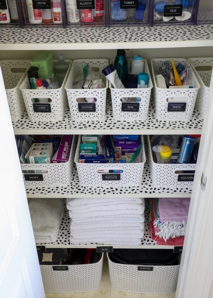 Bathroom closet shelves with white baskets and black labels on top of dotted shelf liner
