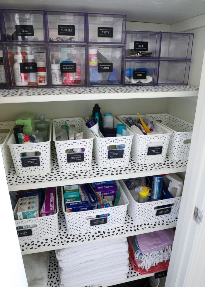 Bathroom closet shelves organized with clear acrylic drawers and white baskets with black labels