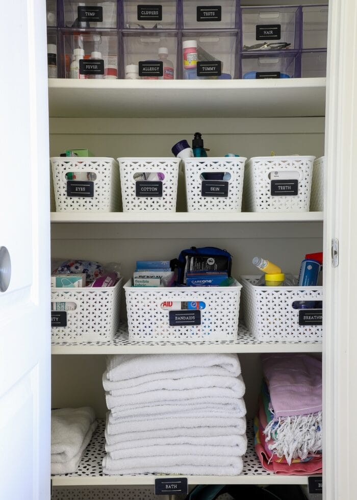 Bathroom closet shelves organized with white baskets and black labels on top of dotted shelf liner