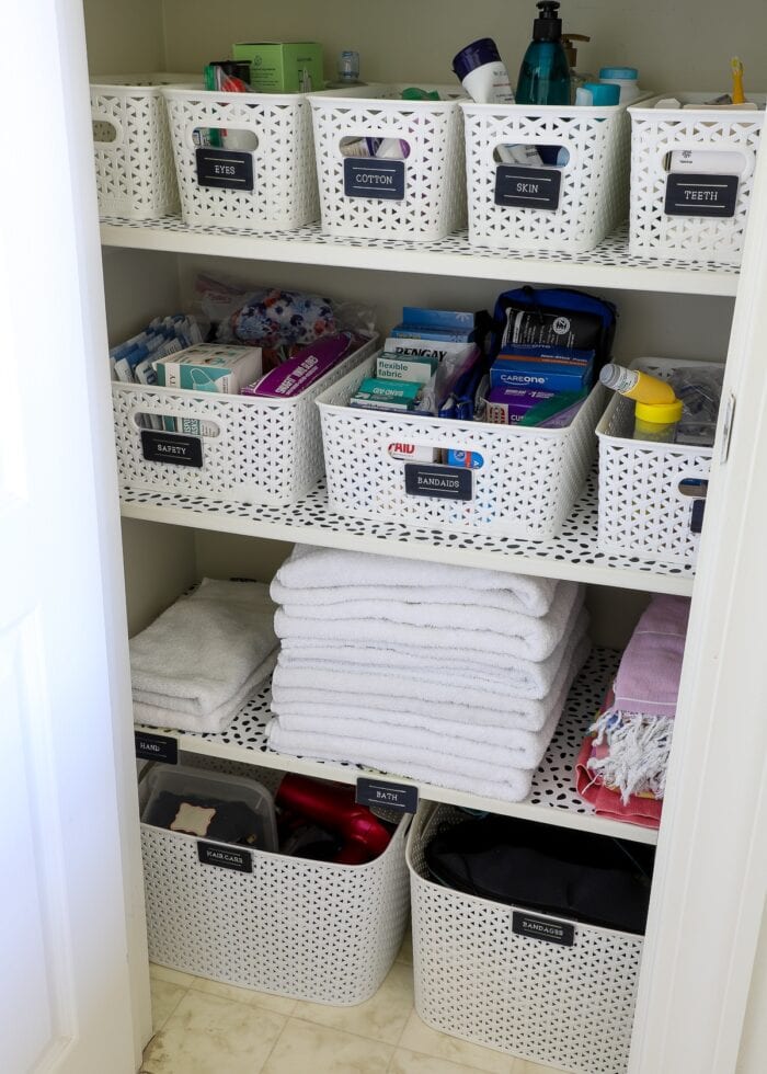 Bathroom closet shelves organized with white baskets and black labels on top of dotted shelf liner