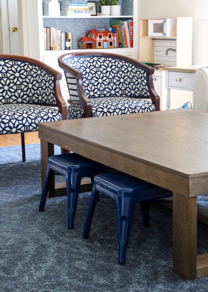 Large wooden coffee table on top of blue rug with two blue metal stools tucker underneath