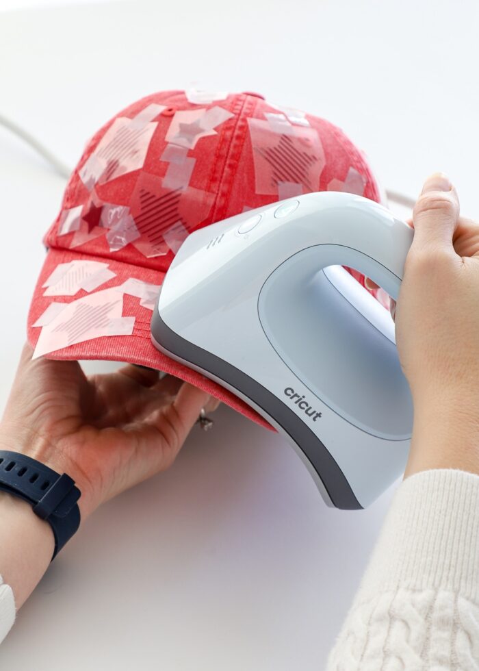 Hand holding Cricut Hat Press on top of red hat with iron-on design all over it