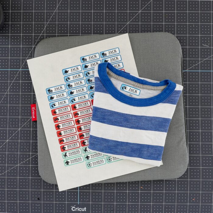 A blue and white striped shirt shown next to a sheet of iron on labels on top of a Cricut cutting mat