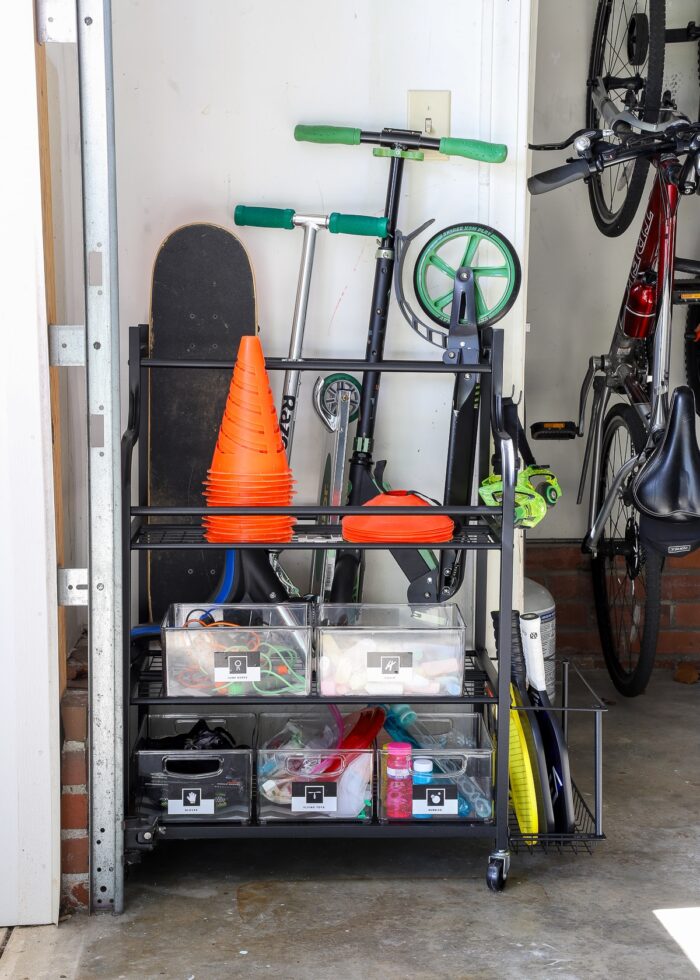 Sports equipment storage rack along a garage wall loaded up with cones, scooters, jump ropes, pickle ball supplies, bubbles, chalk and more.