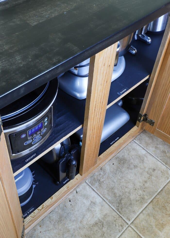 Lower kitchen cabinets holding small appliances with white labels on black shelf liner