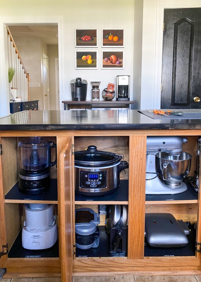 Various small kitchen appliances lined up inside lower kitchen cabinets under a kitchen island.