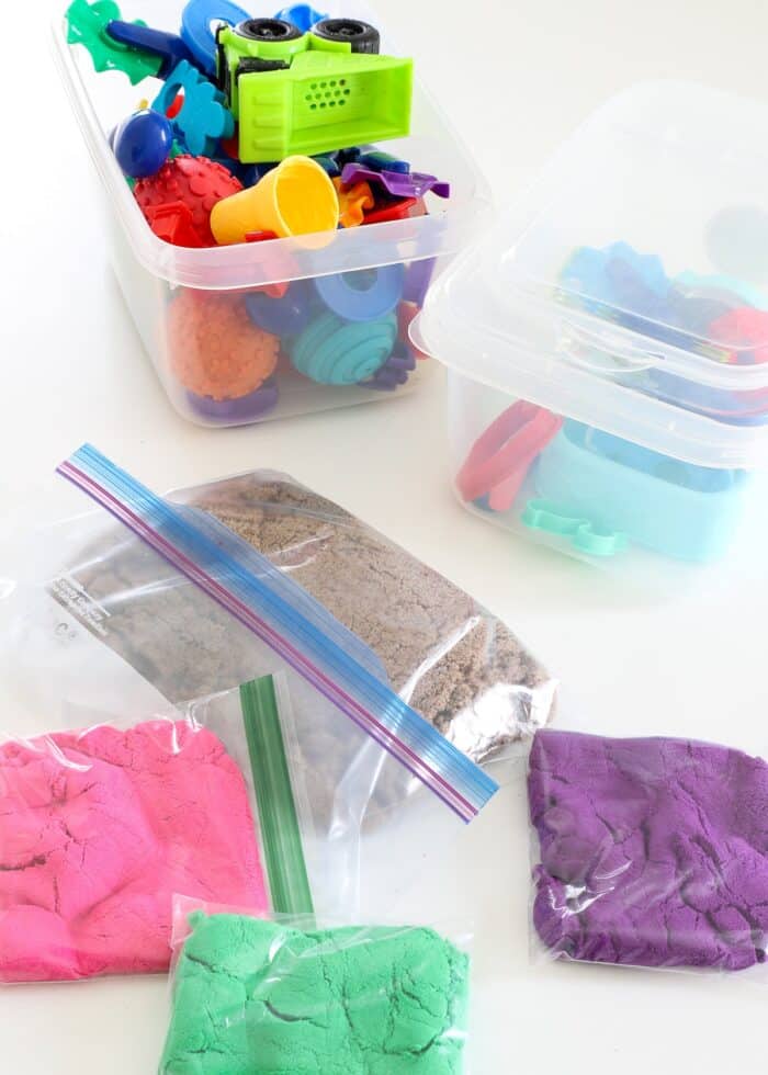 Different colors of kinetic sand in plastic bags alongside bins of sand toys and tools