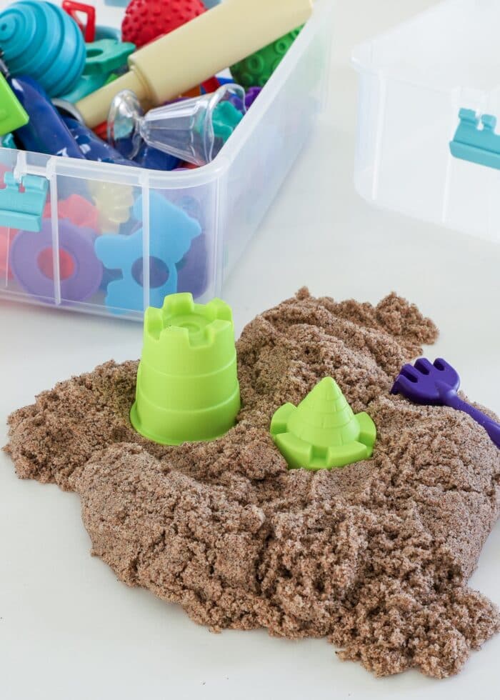 A pile of Kinetic sand on a white table shown alongside a bin of toys and tools