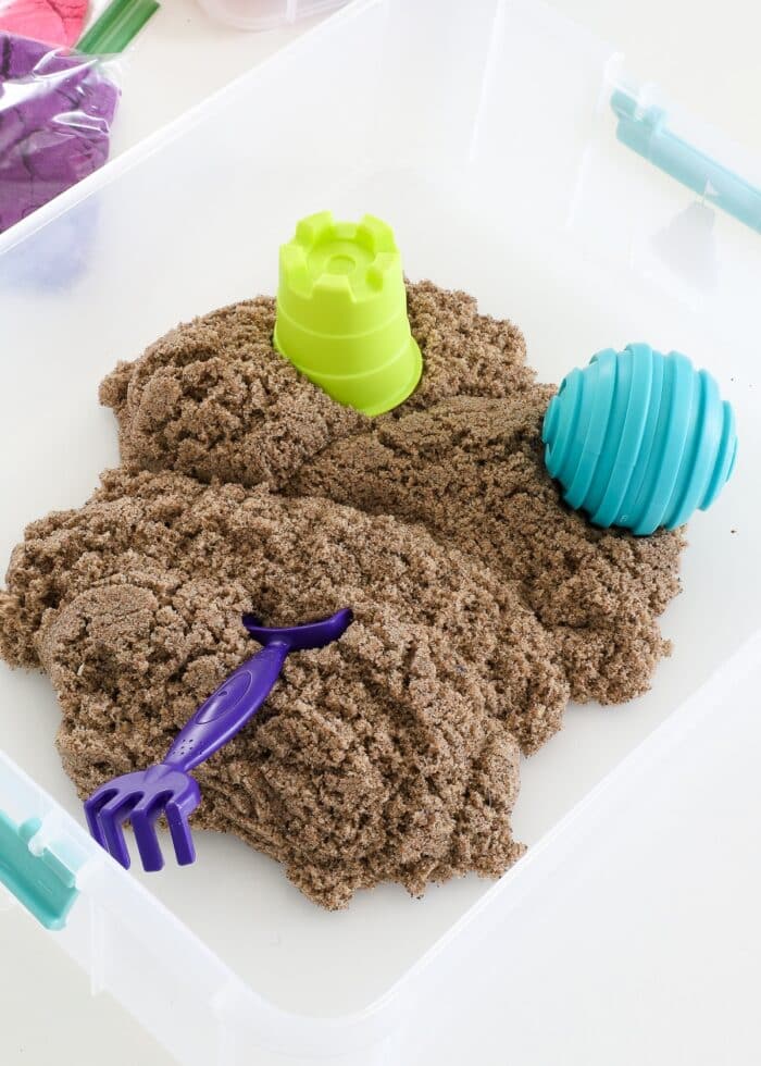 A pile of Kinetic sand inside a clear bin shown with sand toys and tools