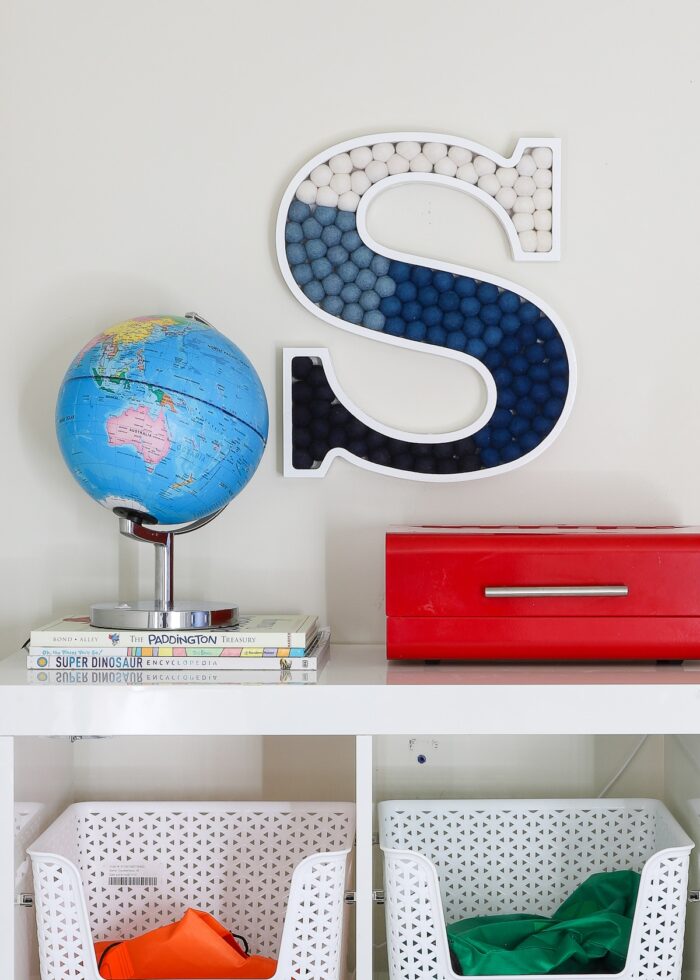 A wall letter "S" filled with blue pom poms hung on a wall above a white bookshelf