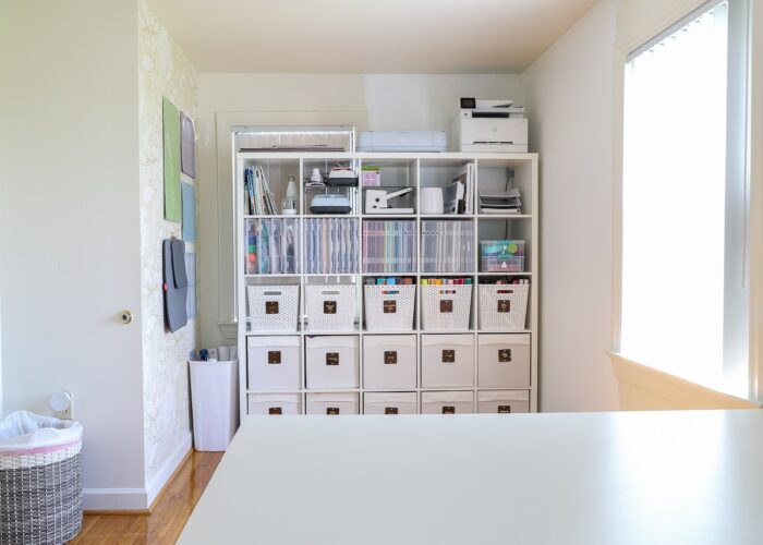 White IKEA Kallax unit on tucked into a nook of a craft room, holding a variety of bins and baskets filled with craft supplies