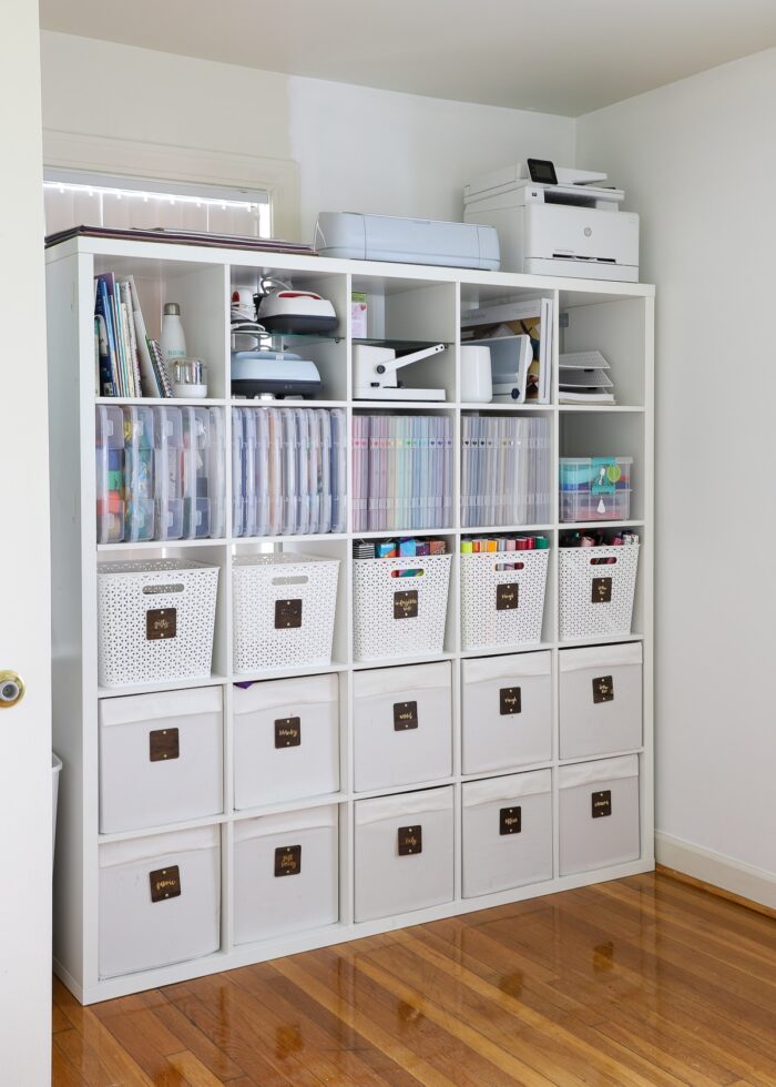 White IKEA Kallax unit on tucked into a nook of a craft room, holding a variety of bins and baskets filled with craft supplies