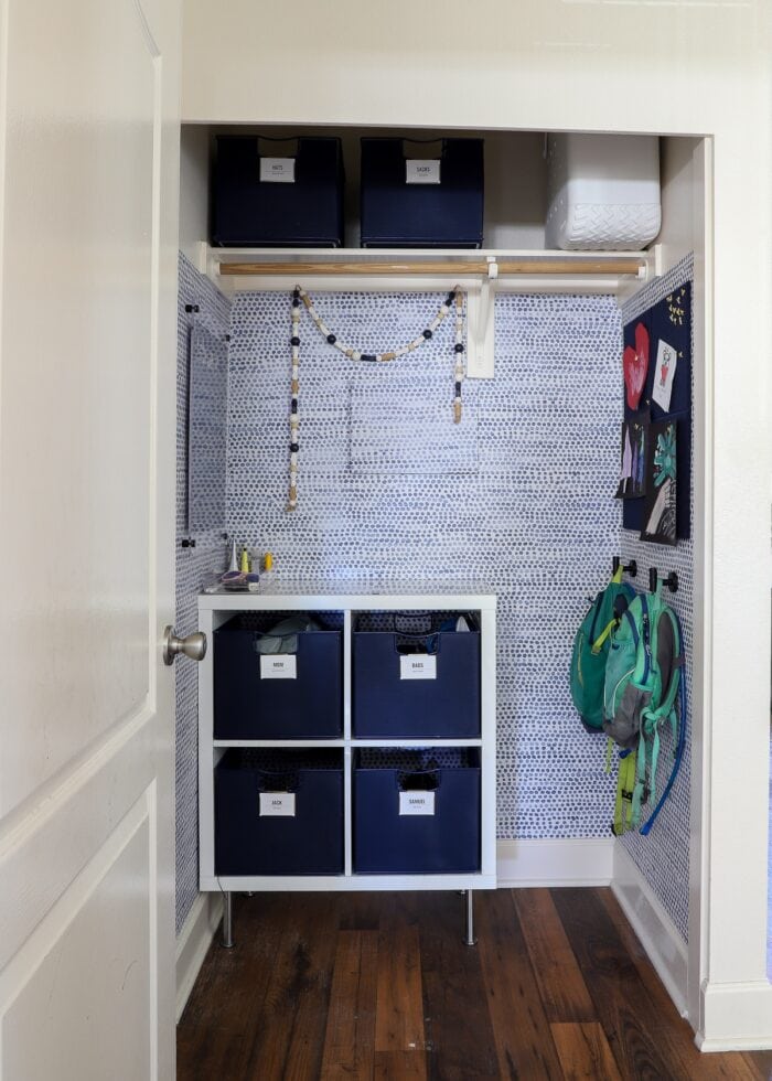 A closet turned into a cute mudroom with blue and white wallpaper, dry-erase calendar and blue baskets inside a white cubby shelf