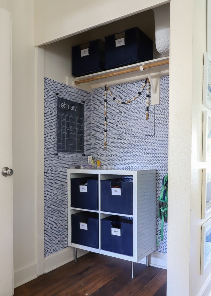 A closet turned into a cute mudroom with blue and white wallpaper, dry-erase calendar and blue baskets inside a white cubby shelf