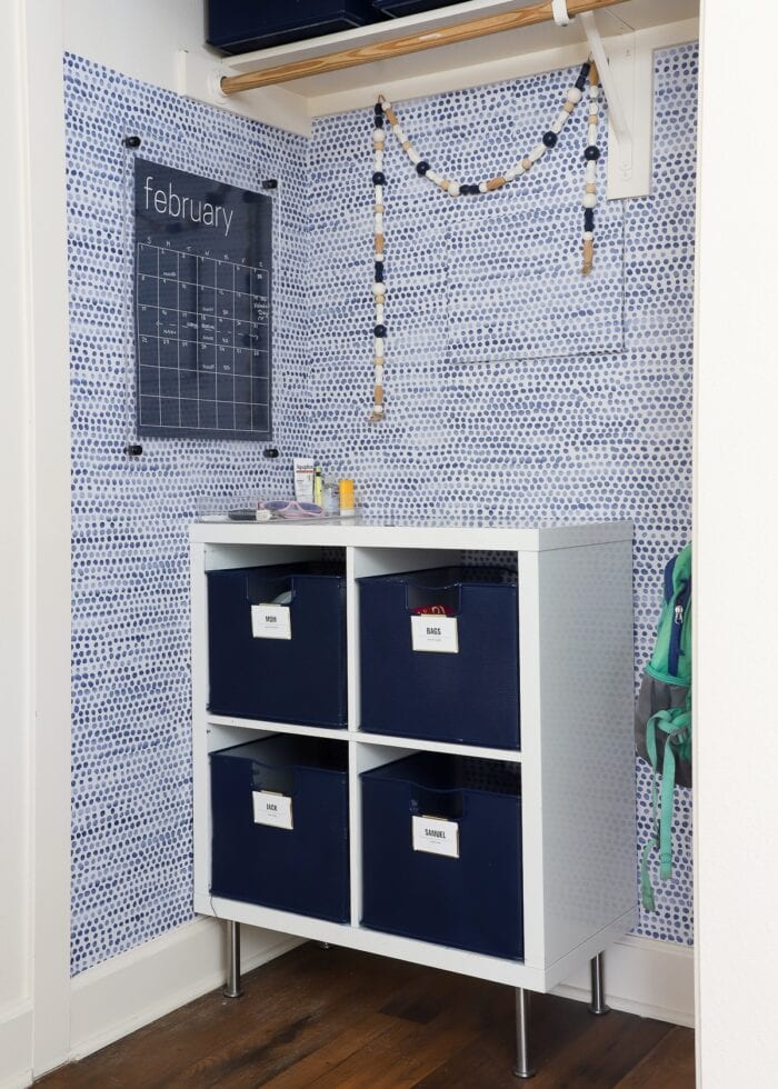 A closet turned into a cute mudroom with blue and white wallpaper, dry-erase calendar and blue baskets inside a white cubby shelf