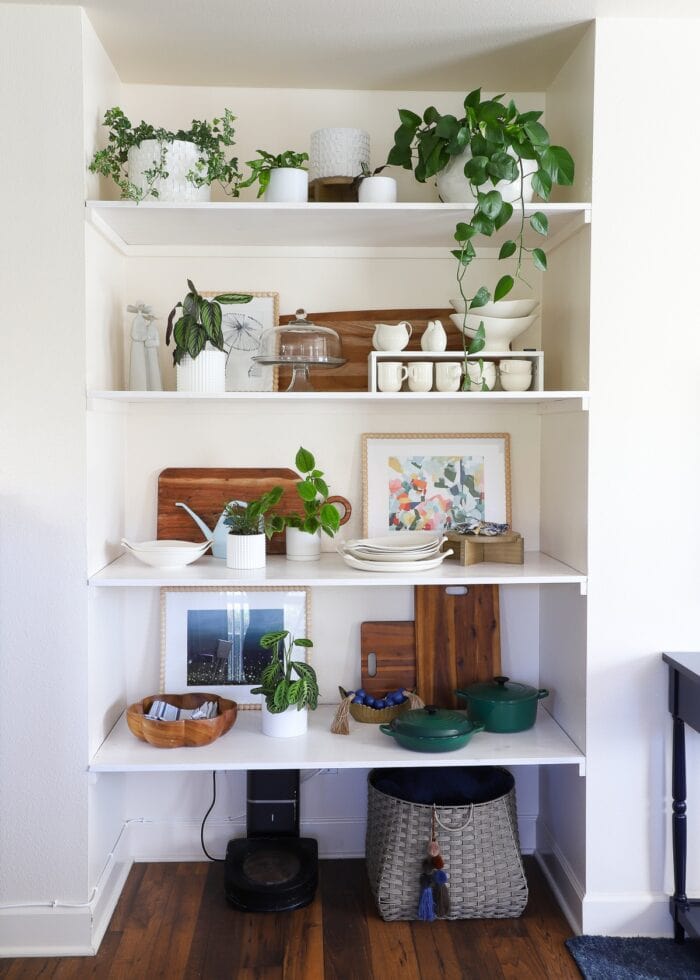 A floor-to-ceiling wall nook outfitted with basic wooden floating shelves that hold dishes, plants, artwork, and decor accessories.