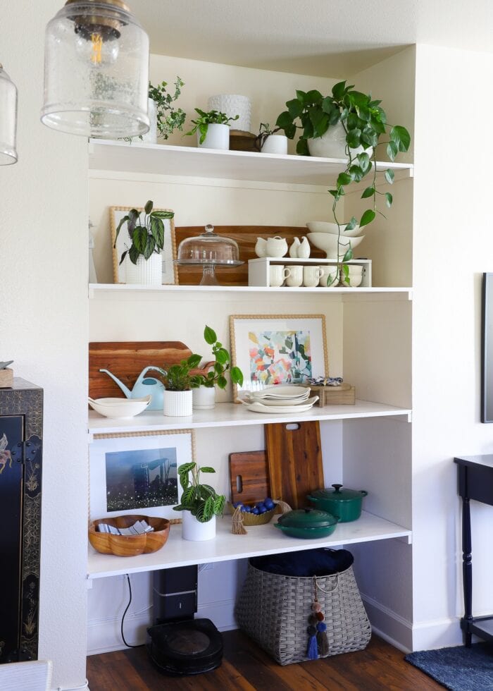 A floor-to-ceiling wall nook outfitted with basic wooden floating shelves that hold dishes, plants, artwork, and decor accessories.