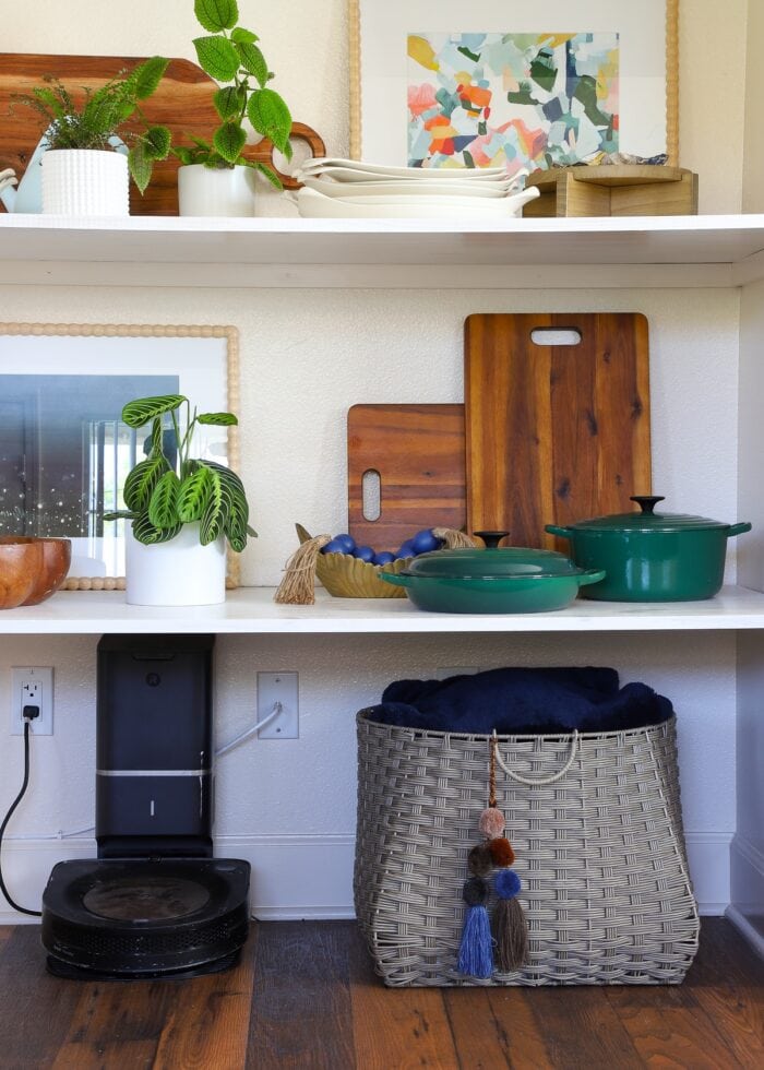 A basket full of blankets and a robot vacuum tucked under a series of floating shelves built into an open nook