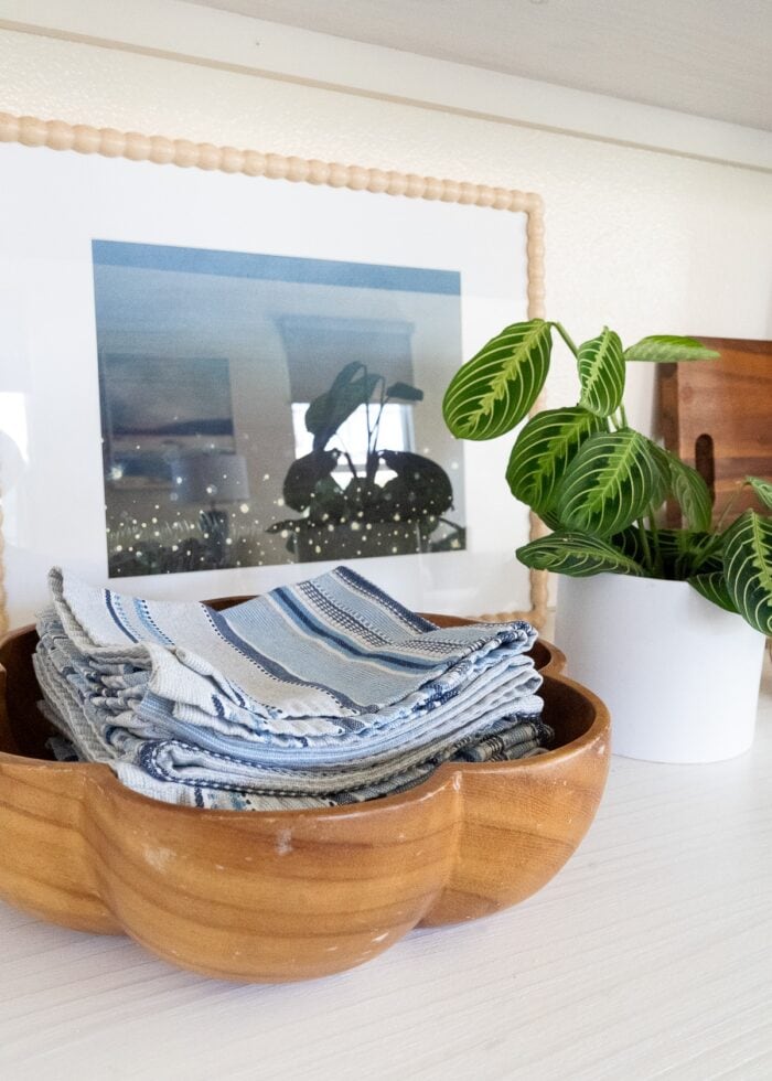 A wooden bowl full of cloth napkins alongside some artwork and a plant