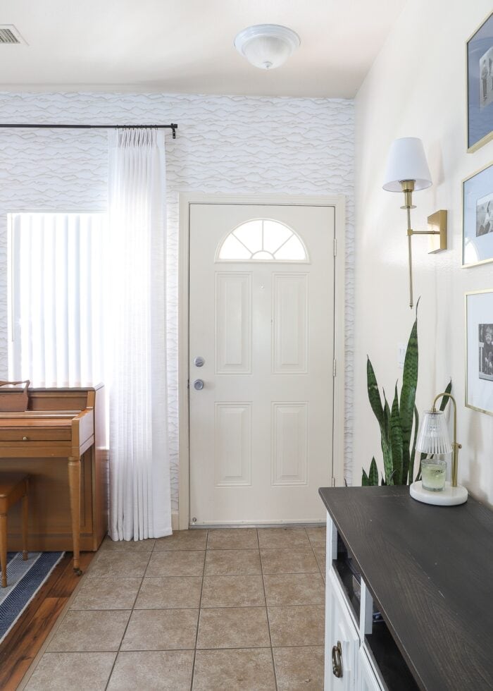 the interior shot of a foyer with beige front door, white and cream wallpaper and sconces on the wall