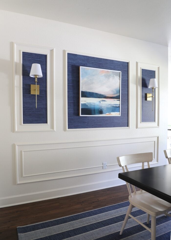 An ivory wall filled with with trim frames, blue grasscloth wallpaper, a single piece of artwork and gold wall sconces