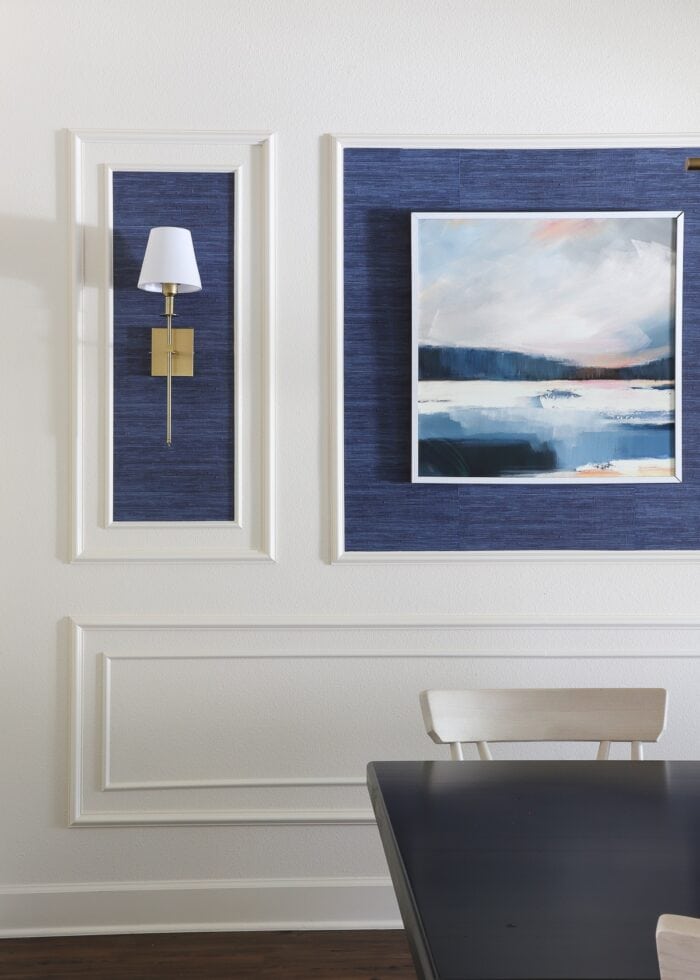 An ivory wall filled with with trim frames, blue grasscloth wallpaper, a single piece of artwork and gold wall sconces