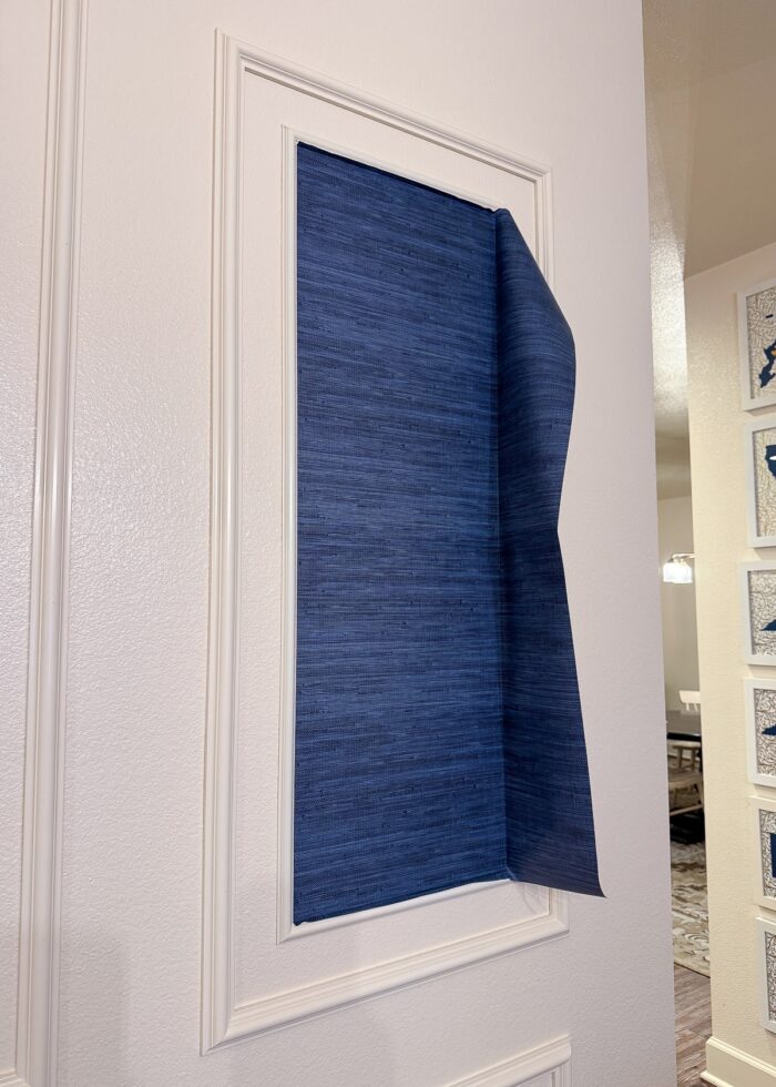 A blue grasscloth wallpaper scrap inside a trim frame on a wall