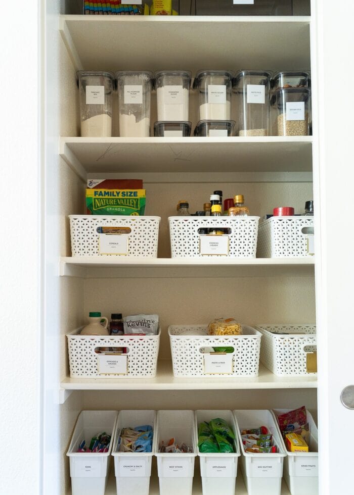 An organized pantry with all white bins, baskets, and clear Rubbermaid Brilliance Pantry Canisters with big white labels