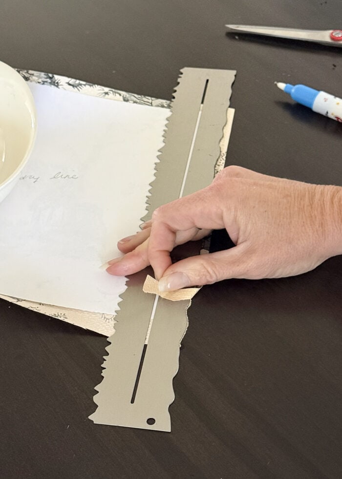 A hand tearing off the edge of a scrap of vintage wallpaper using a tearing ruler
