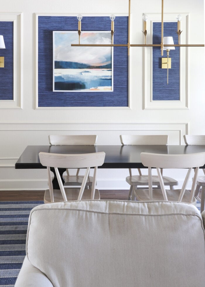 A dark wood dining table with light colored wood dining chairs against a cream wall with blue artwork.
