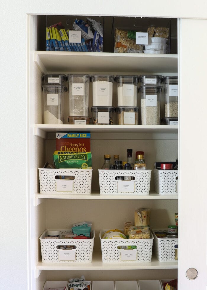 An organized pantry with bins of backstock items on the top shelf